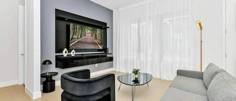 Una sala de estar moderna con un televisor de pantalla plana montado en la pared, un sofá gris, un sillón negro decorativo, una pequeña mesa redonda de centro y una lámpara de pie.