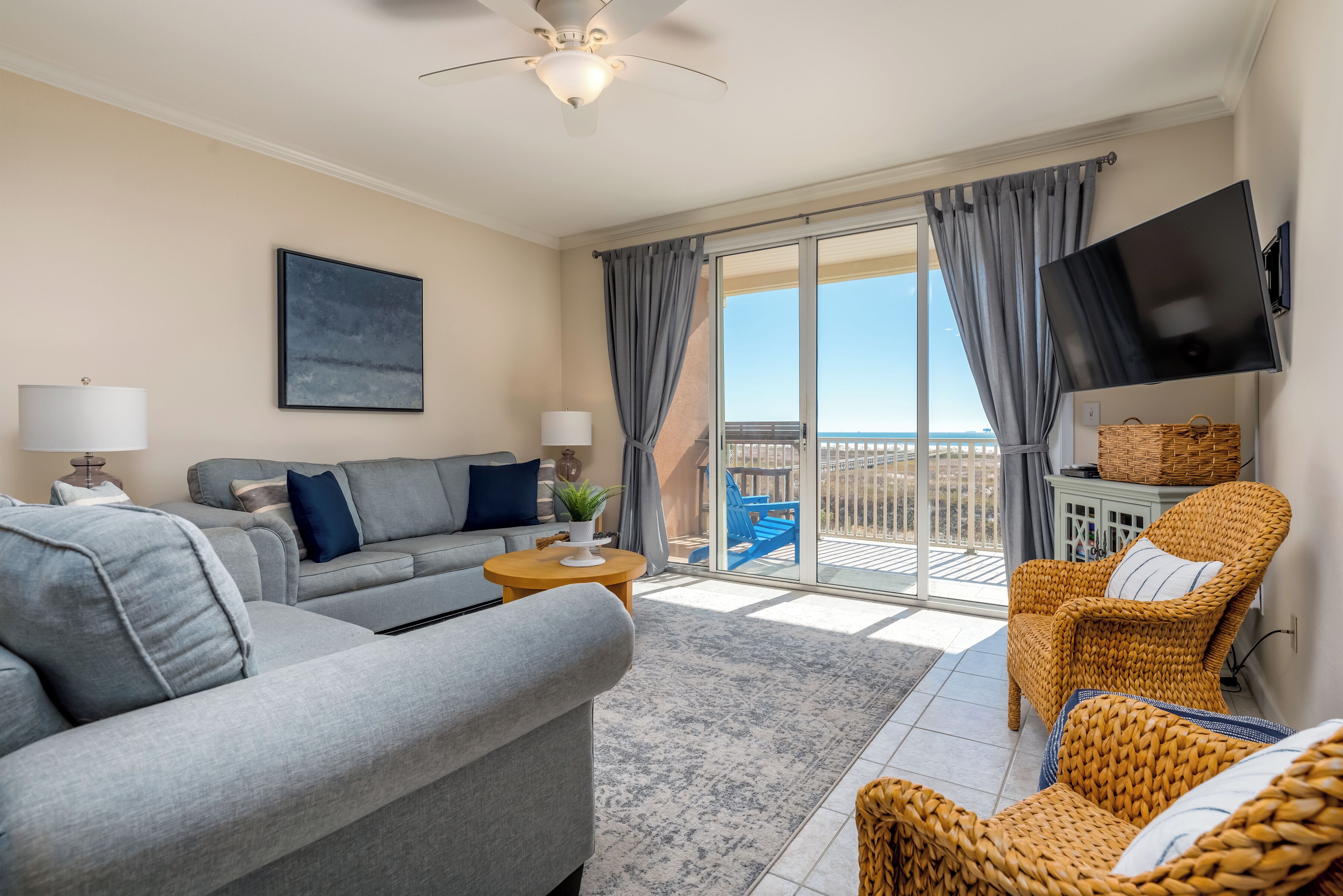 A modern living room with a grey sofa, a wicker chair, a flat-screen TV, and a balcony with a view.