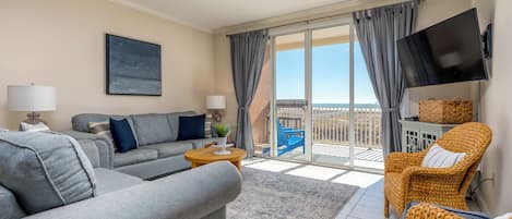 A modern living room with a grey sofa, a wicker chair, a flat-screen TV, and a balcony with a view.