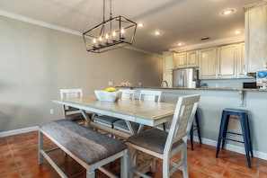 A dining area with a wooden table and chairs, a chandelier, and a kitchen in the background. - Side by Side Beach Condos | Sleeps 18, Pools + Beach Access (Dauphin Island)