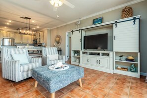 A modern living room with a sofa, ottoman, TV, and entertainment unit. - Side by Side Beach Condos | Sleeps 18, Pools + Beach Access (Dauphin Island)