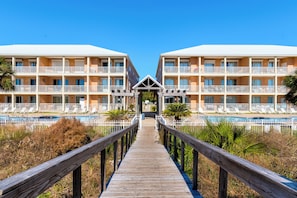 Un resort con un paseo marítimo de madera que conduce a un área de piscina, rodeado de palmeras y un cielo azul claro.