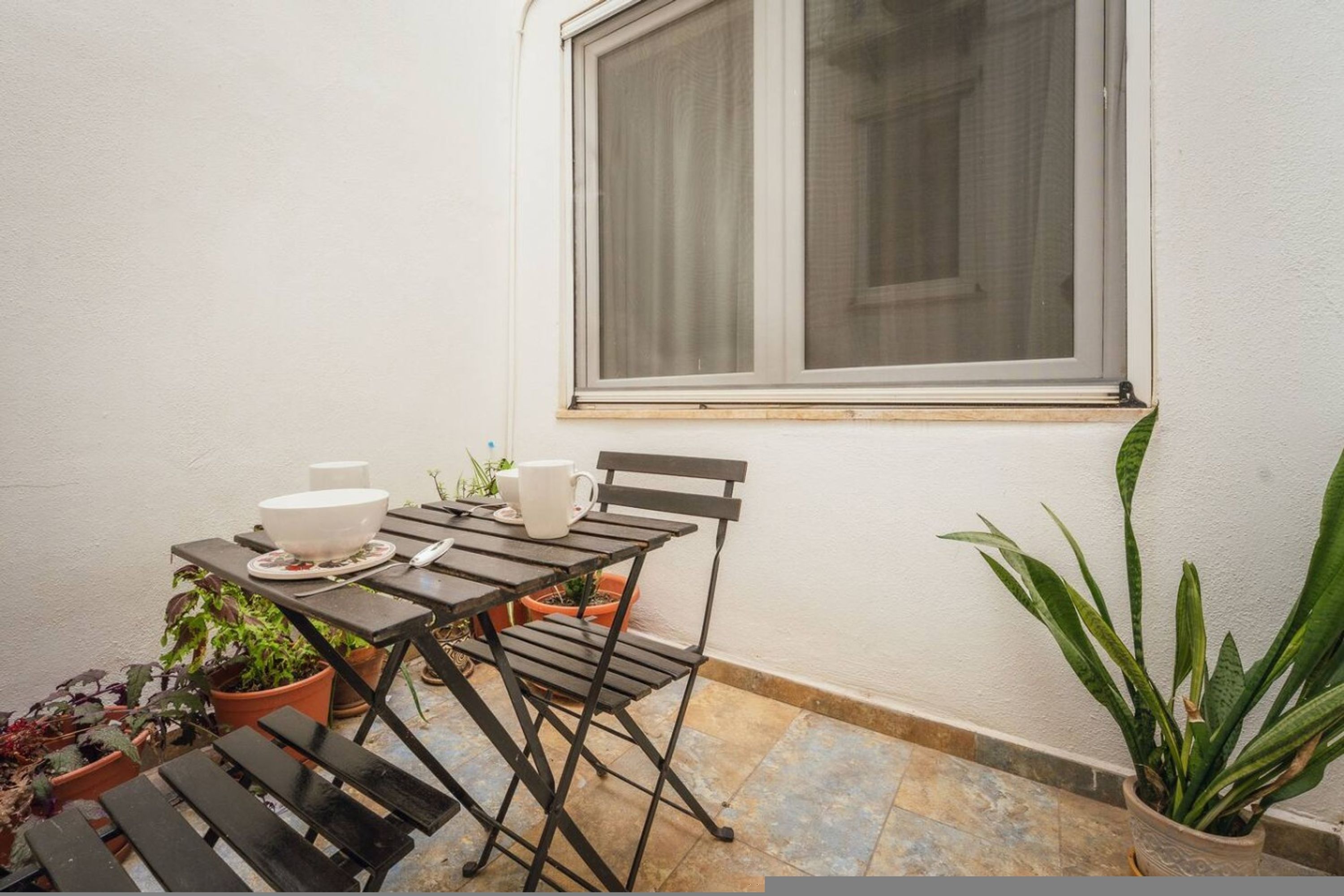 A small outdoor seating area with a table and chairs, a window, and potted plants.