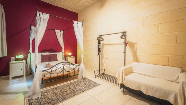 A bedroom with a four-poster bed, a nightstand with a lamp, a bench, and a wall-mounted drying rack. - Casa Due Balli 400-year Historic Valletta House (Valletta)