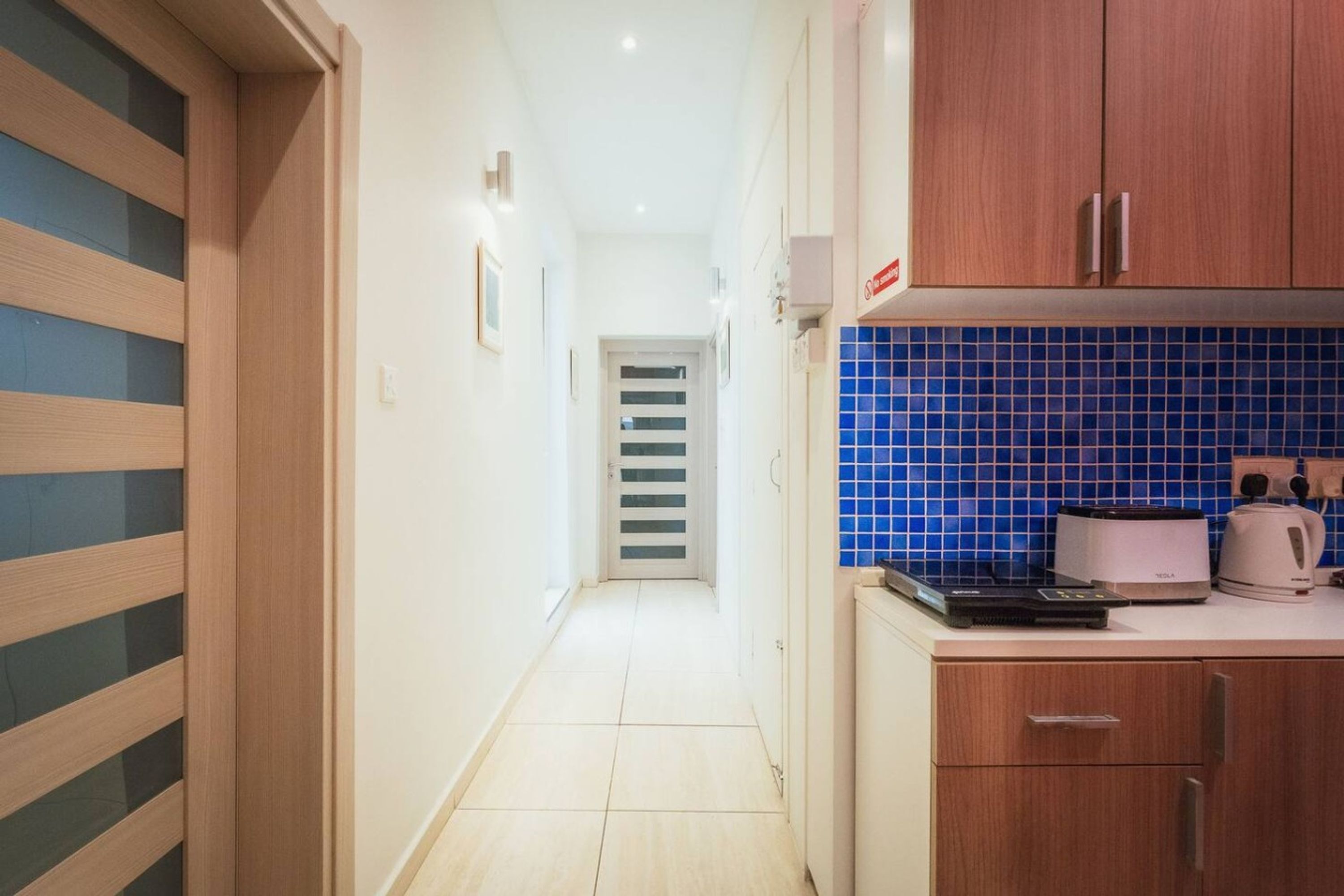 Un corridoio stretto che conduce a una porta con decorazioni a righe, un angolo cottura con piastrelle blu e un piano di lavoro con un tostapane e un bollitore.