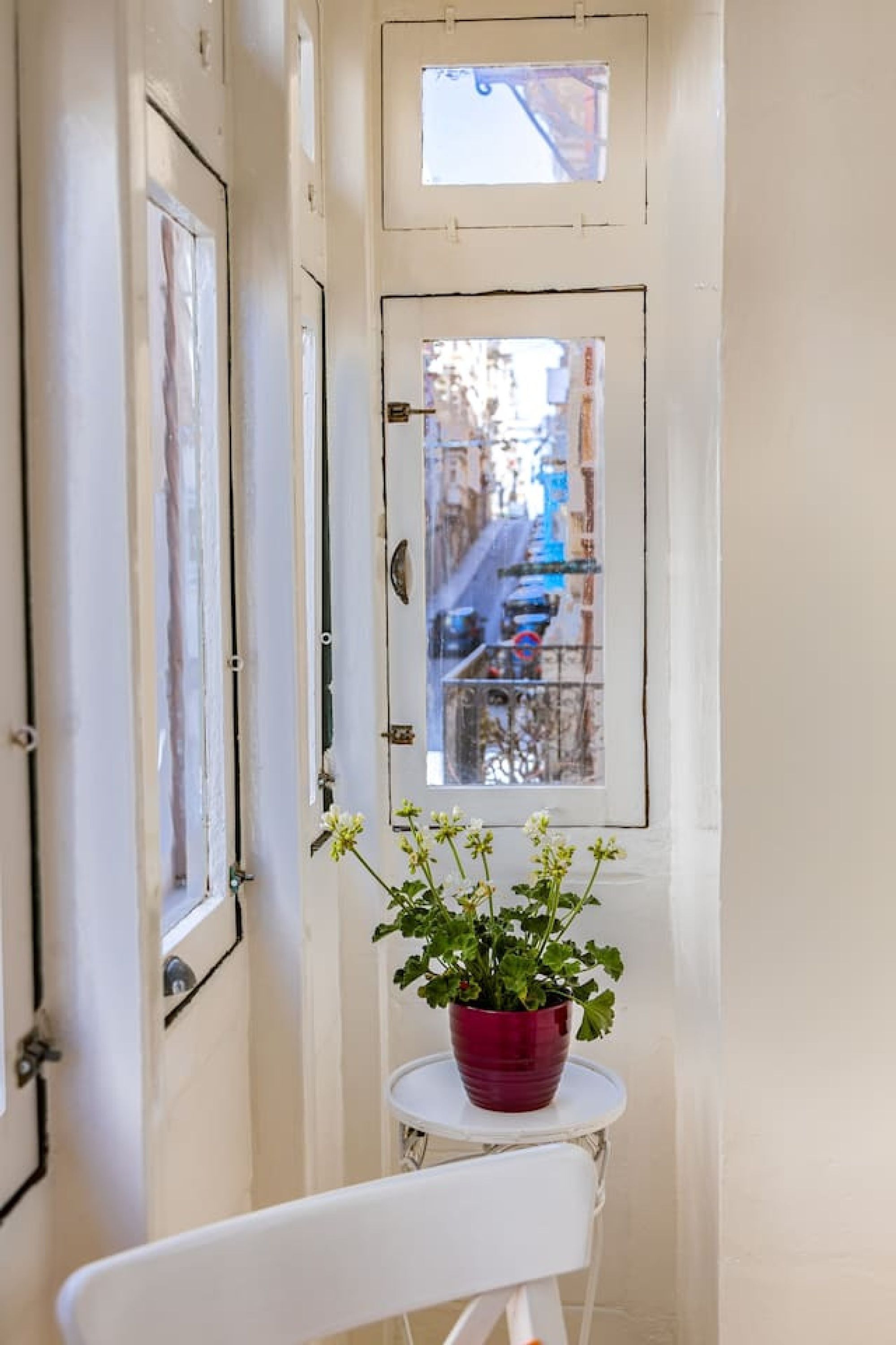 A narrow hallway with a white door and a window with a view of a street. A red pot with green plants is placed on a small white table.
