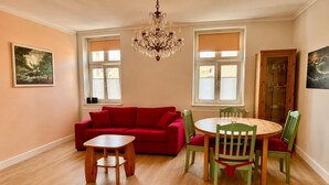 Una habitación con un sofá rojo, una mesa de madera, sillas verdes, una lámpara de araña y dos ventanas con persianas.