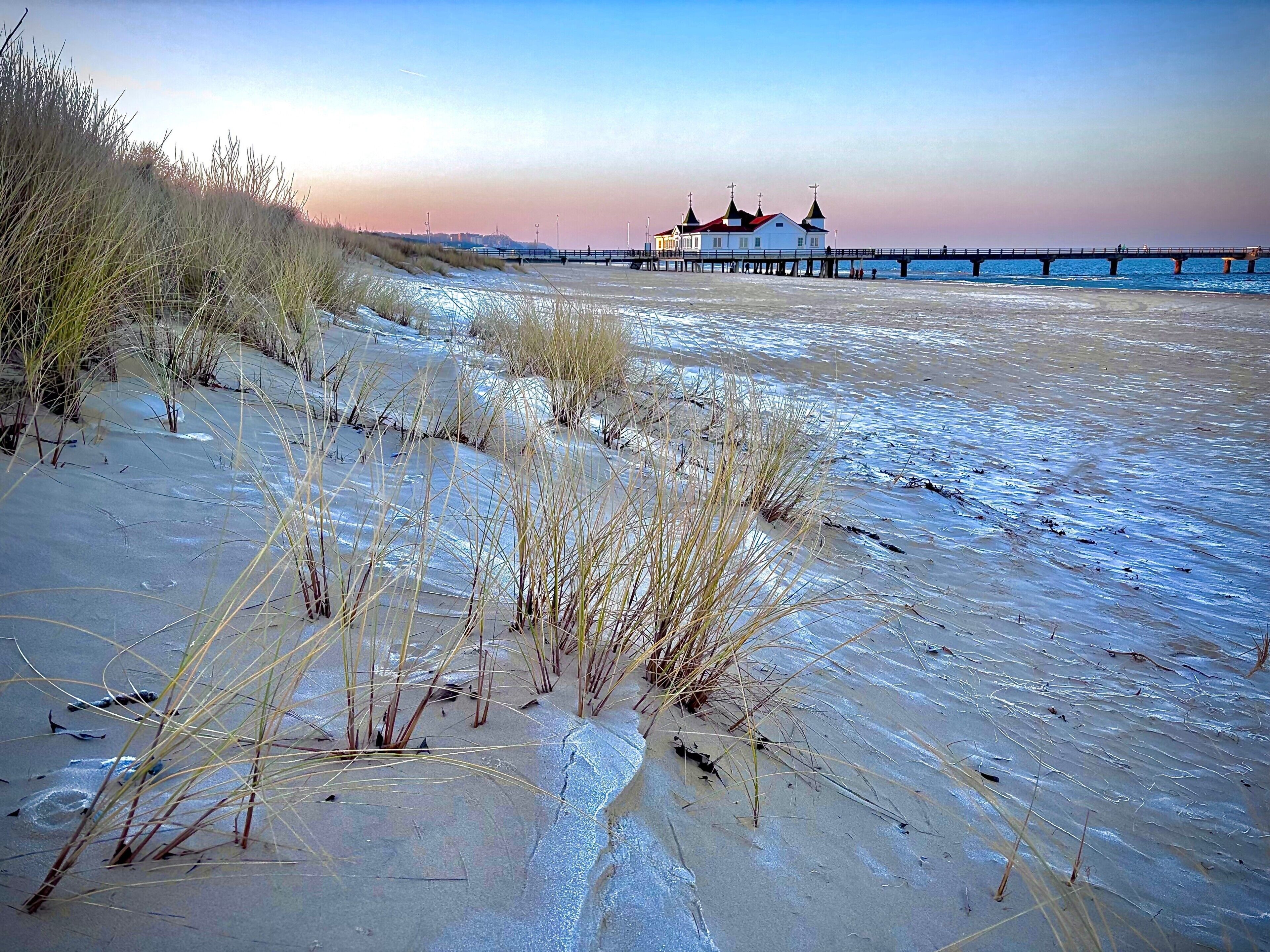 Ein Strand mit einer Mole, die ins Wasser ragt, ein Gebäude mit rotem Dach und üppiger Vegetation.