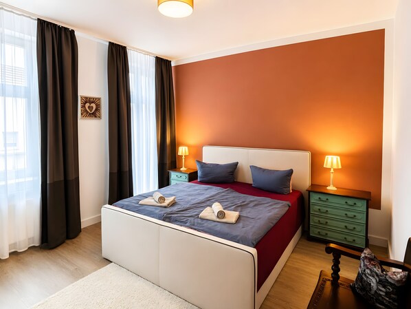 A bedroom with a bed, two bedside tables, a nightstand, a window with curtains, and a wall with a warm color.