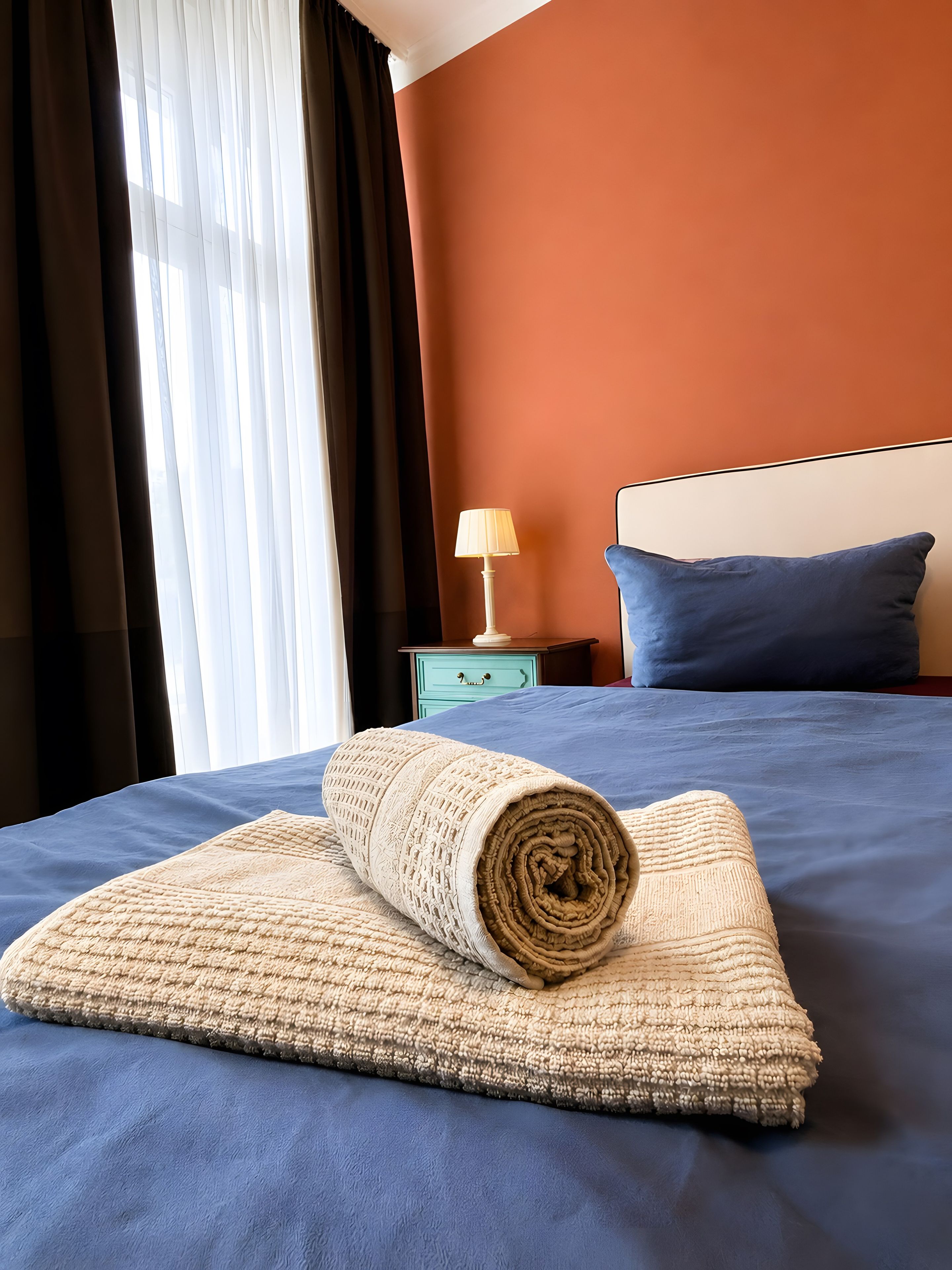 Ein ordentlich bezogenes Bett mit einer zusammengerollten Decke, einer Nachttischlampe und einem blauen Kissen auf einer blauen Bettdecke.