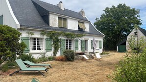 Uma casa com telhado inclinado, varanda da frente com mesa e cadeiras, e um pequeno galpão ao fundo.