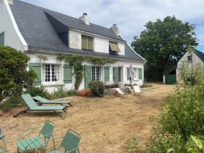 Ein Haus mit geneigtem Dach, einer Veranda mit Tisch und Stühlen sowie ein kleines Gartenhaus im Hintergrund.