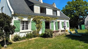 Uma casa de dois andares com telhado inclinado, persianas verdes e um jardim com um banco e mobiliário de exterior.