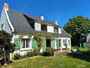 Ein zweigeschossiges Haus mit geneigtem Dach, grünen Fensterläden und einem Garten mit Bank und Gartenmöbeln.