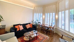 A living room with a sofa, coffee table, and dining area with chairs. There are large windows with blinds, a ceiling light fixture, and a plant. - Beautiful and spacious 2 Double Bed, 2 Bath Garden Flat, Ealing, London, W5 (London)