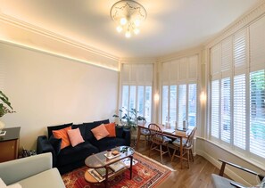 A living room with a sofa, coffee table, and dining area with chairs. There are large windows with blinds, a ceiling light fixture, and a plant. - Beautiful and spacious 2 Double Bed, 2 Bath Garden Flat, Ealing, London, W5 (London)