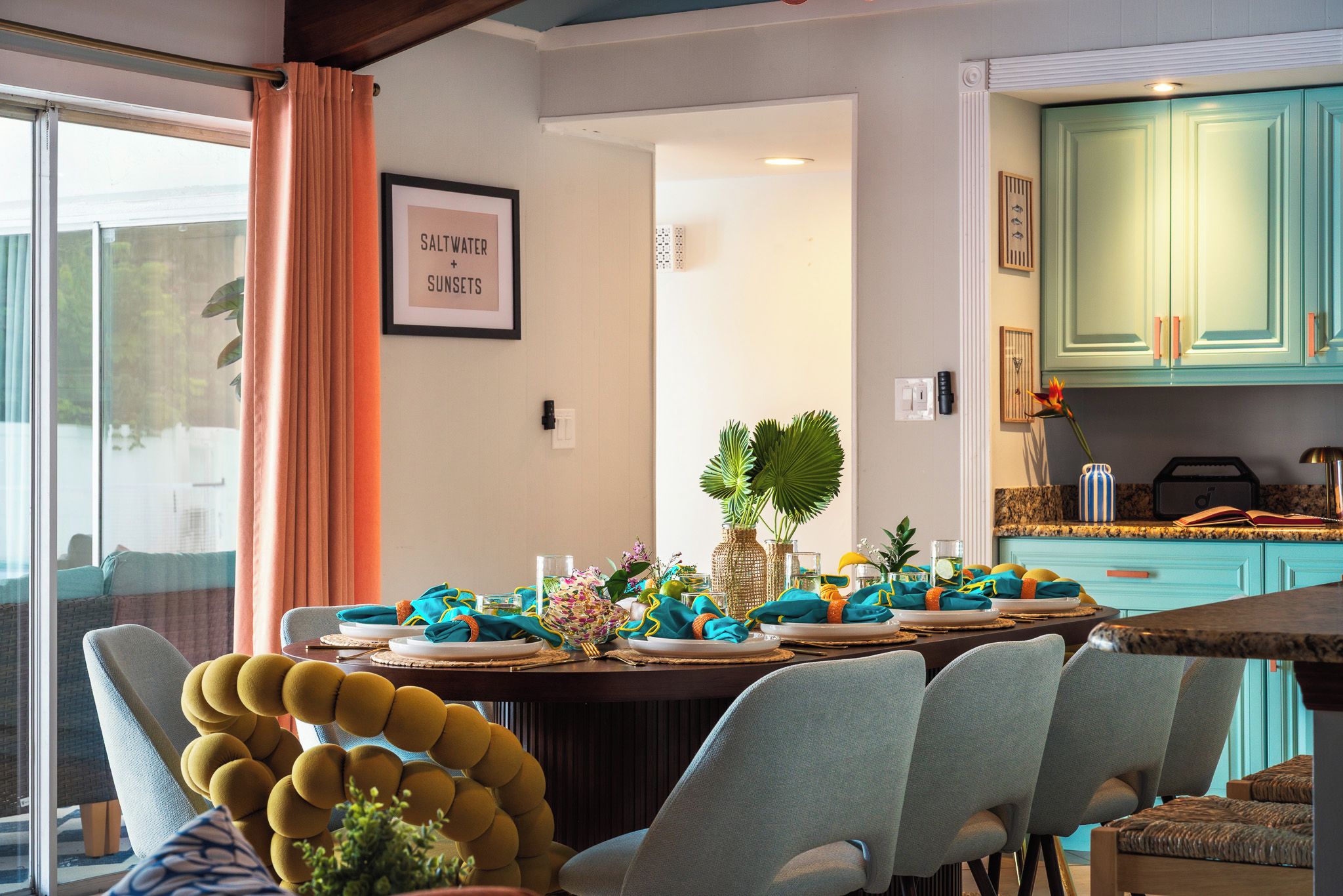 A dining area with a table set for four, chairs, a plant, and a framed picture on the wall.