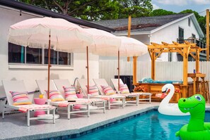 Un espace aménagé au bord de la piscine, avec des chaises longues et des parasols, une pergola en bois, ainsi qu’un flotteur gonflable en forme de cygne et de crocodile.