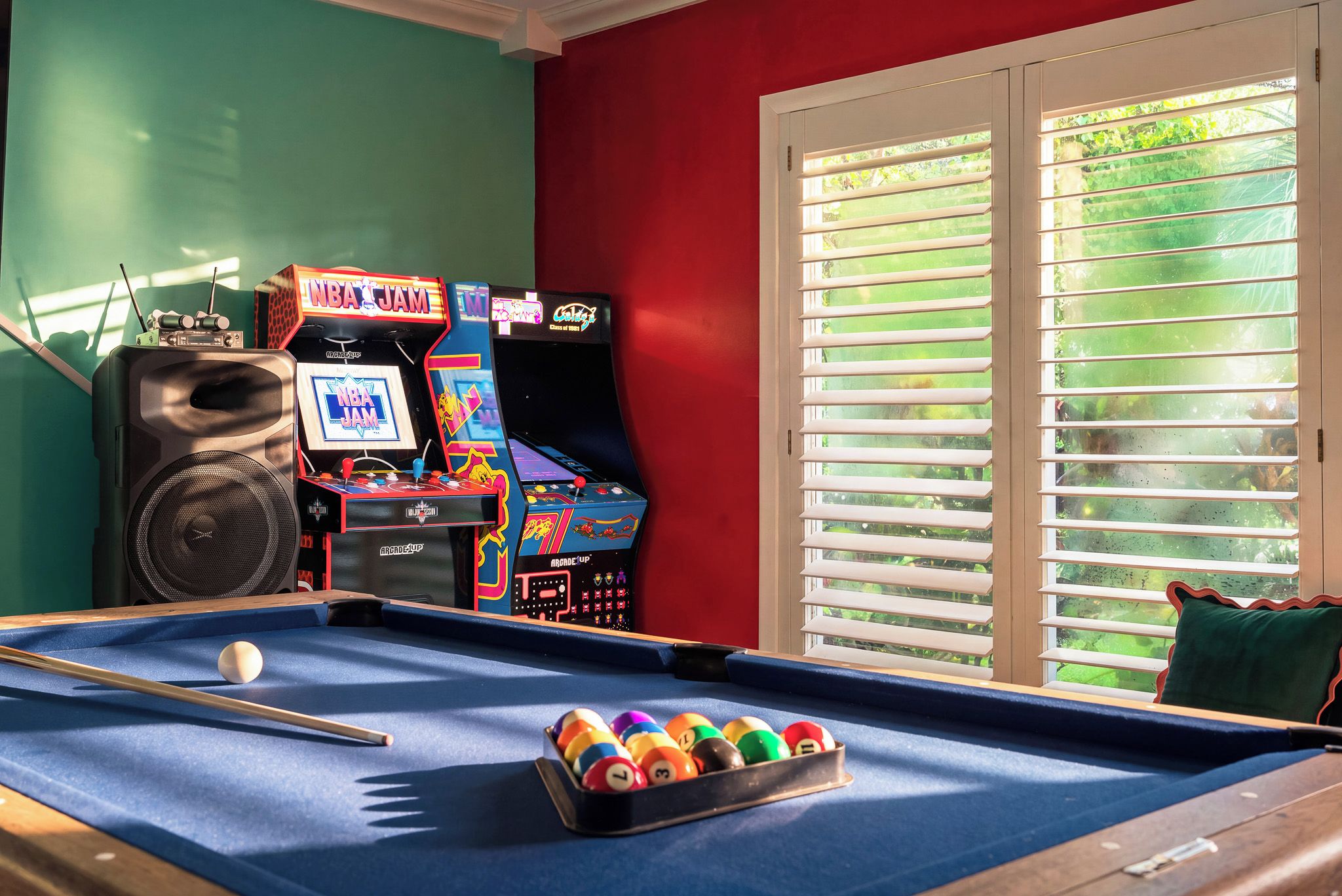 Una mesa de billar con taco y bolas, máquinas de arcade y un altavoz en una habitación con ventana y persianas.