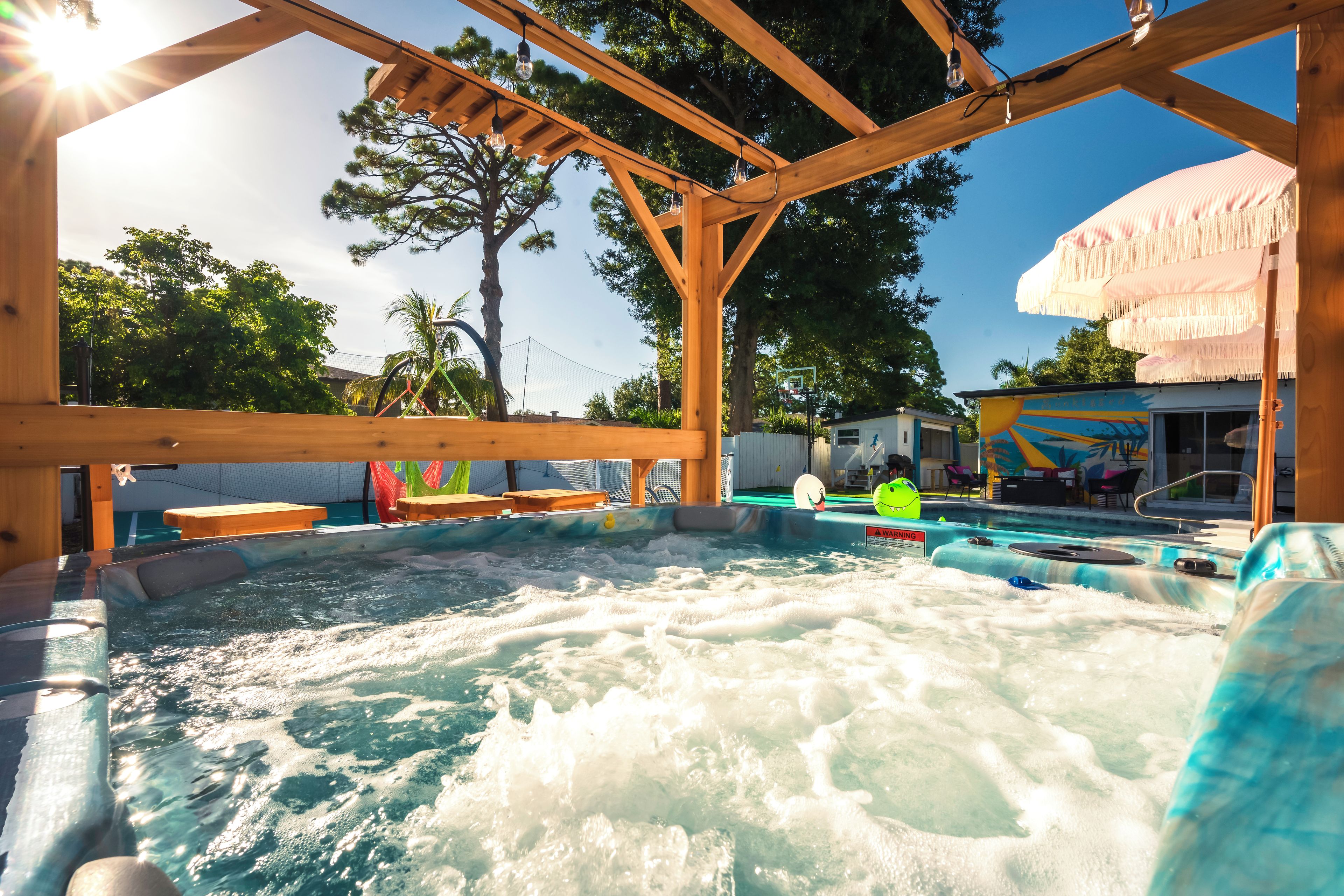 A hot tub area with wooden beams overhead, a pool with a safety sign, and a mural on the building.