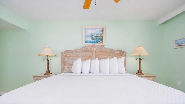A bedroom with a large bed, two bedside tables with lamps, a wooden headboard, and a painting on the wall. - Phoenix Iii 3036 2 Bedroom Condo (Orange Beach)