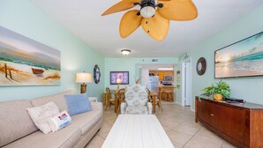 A living room with a sofa, coffee table, and a ceiling fan. There is a dining area with chairs and a table. The room has a kitchen in the background. - Phoenix Iii 3036 2 Bedroom Condo (Orange Beach)