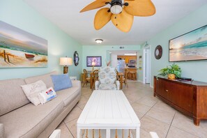 A living room with a sofa, coffee table, and a ceiling fan. There is a dining area with chairs and a table. The room has a kitchen in the background. - Phoenix Iii 3036 2 Bedroom Condo (Orange Beach)