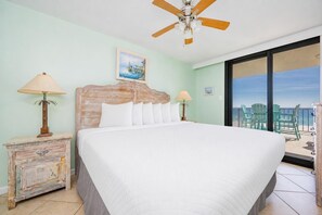 Une chambre d’hôtel avec un lit, un ventilateur de plafond, une tête de lit en bois, une table de chevet, une lampe et une vue sur la plage, visible à travers une grande fenêtre.