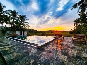 Piscina con terraza de piedra, dos bancos de madera y palmeras.
