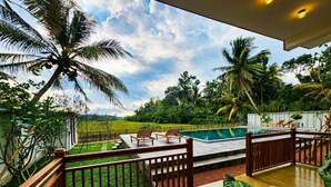 A poolside area with a wooden deck, lounge chairs, and a view of a lush green landscape with palm trees.