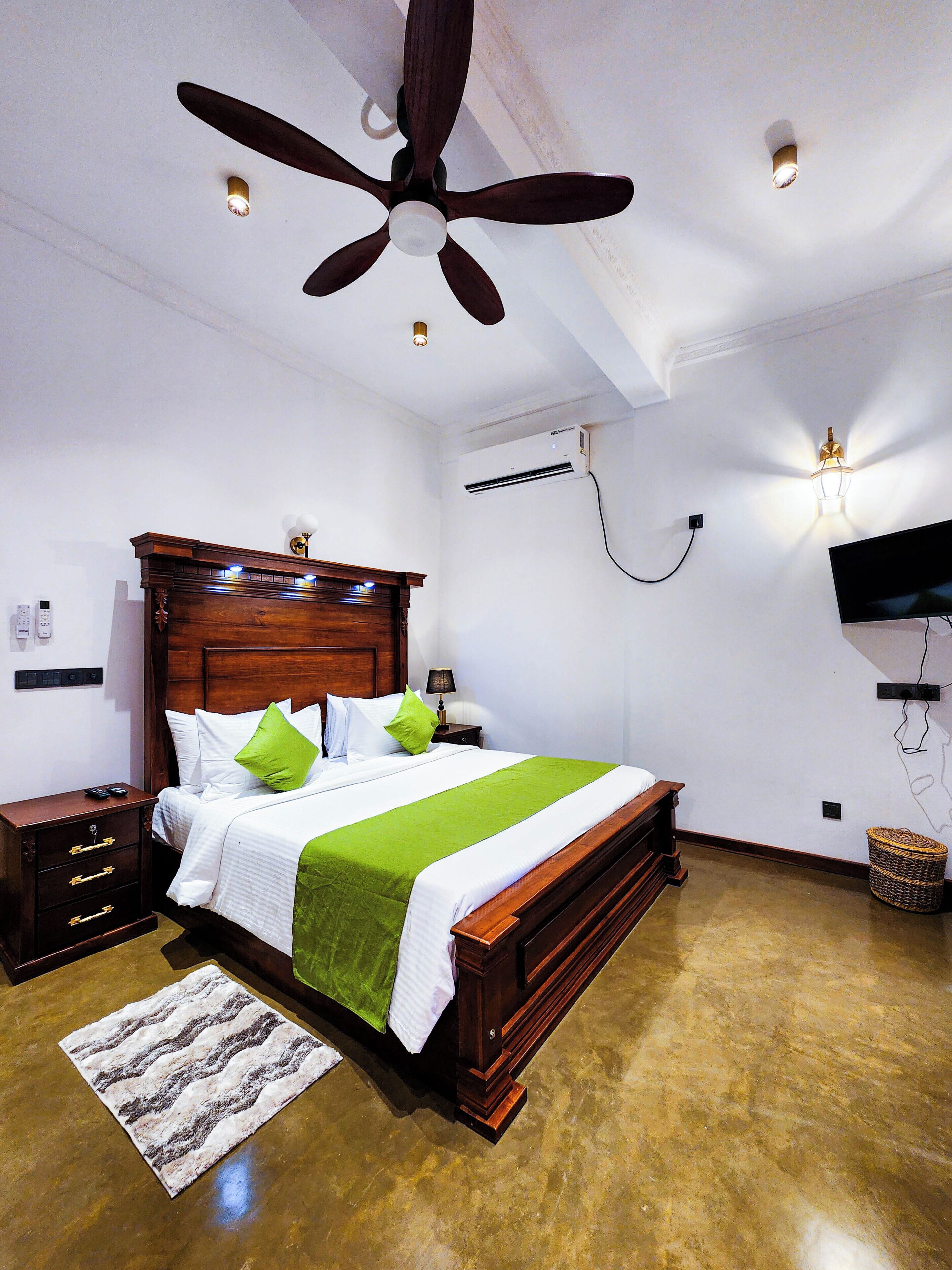 Quarto com cama de madeira, mesas de cabeceira, ventilador de teto, ar condicionado, televisão fixada na parede e tapete estampado no chão.