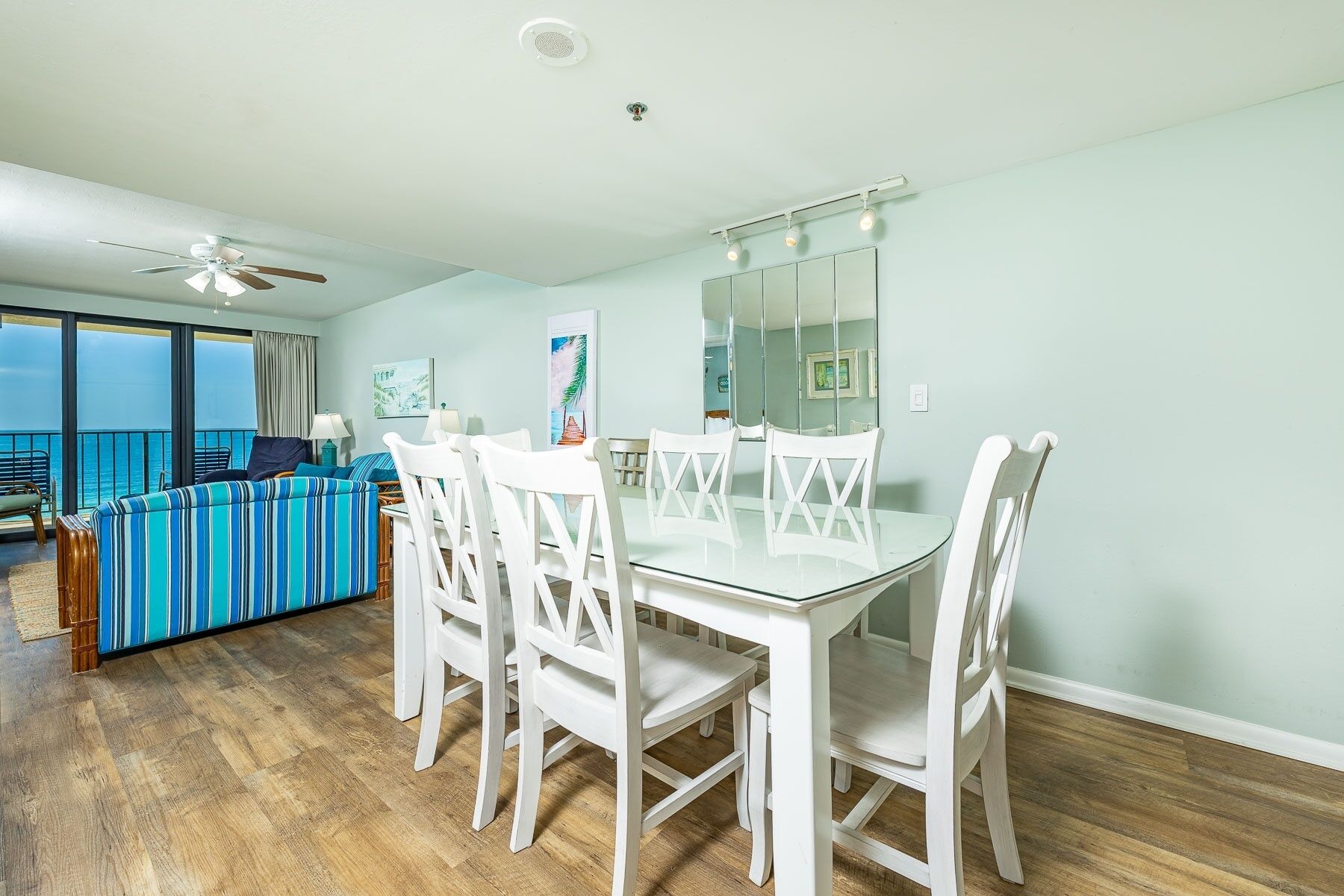 A dining area with a glass table and white chairs, a striped sofa, and a view of the ocean.