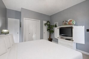 Ein Schlafzimmer mit einem Bett, einem an einem Schrank montierten Fernseher, einer Zimmerpflanze und einer Lampe.