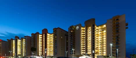 A multi-story hotel with illuminated balconies and a parking lot filled with cars.