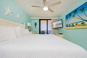 A hotel room with a large bed, a ceiling fan, a TV, and a painting of palm trees. - Phoenix Iii 3128 2 Bedroom Condo (Orange Beach)