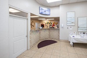 A reception area with a tiled floor, a reception desk, a clock, and a television mounted on the wall. - Phoenix Iii 3128 2 Bedroom Condo (Orange Beach)