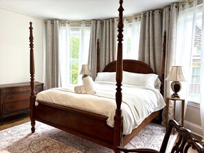 A four-poster bed with a wooden headboard, a bedside table with a lamp, and a view of greenery through the window. - Charming Victorian Duplex 4br Sleeps 10 3 Bedroom Home (Niagara Falls)