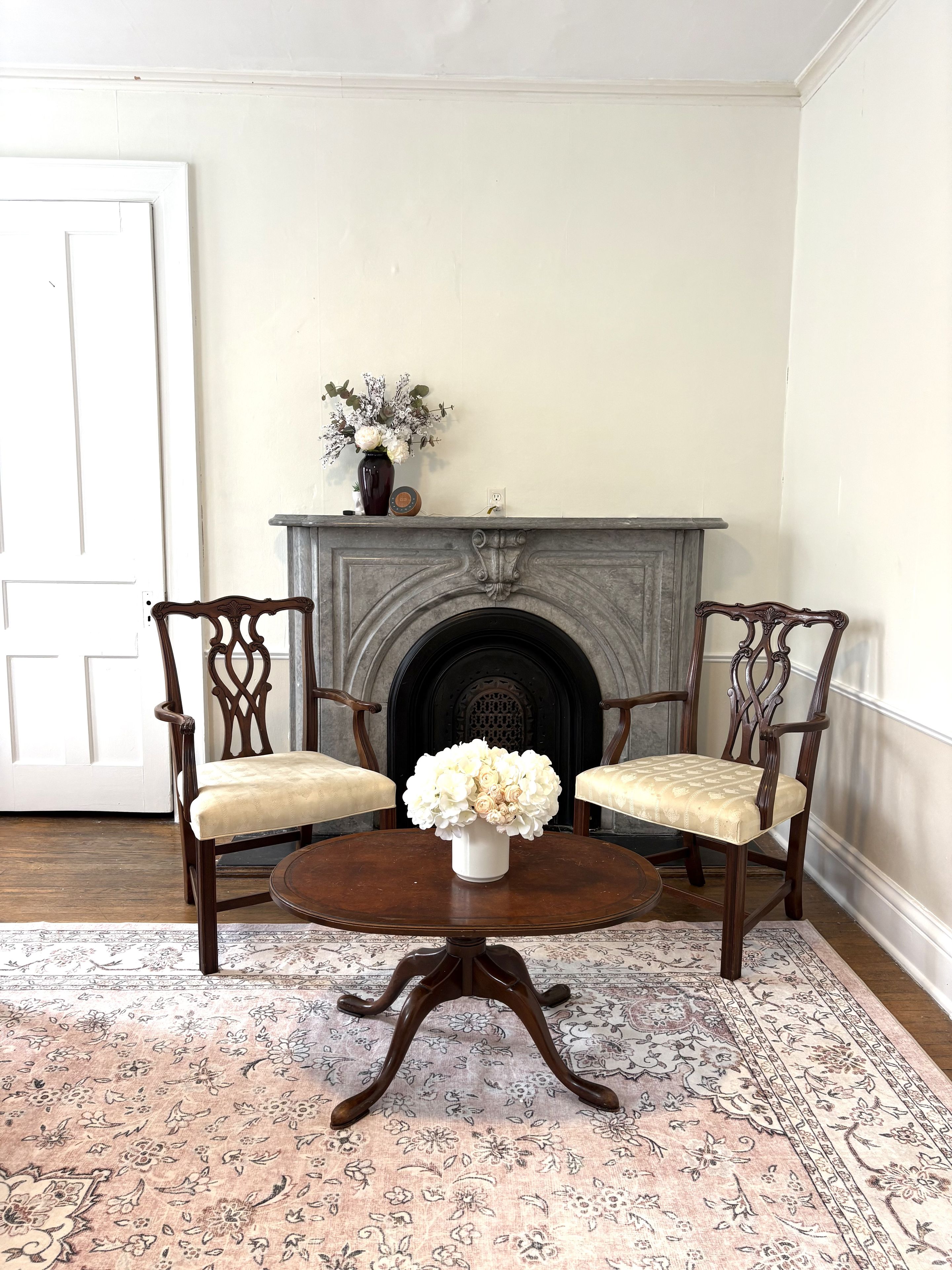 A room with a fireplace, two chairs, a table, and a floral arrangement.
