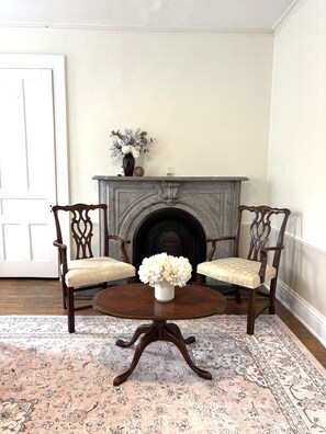 A room with a fireplace, two chairs, a table, and a floral arrangement. - Charming Victorian Duplex 4br Sleeps 10 3 Bedroom Home (Niagara Falls)