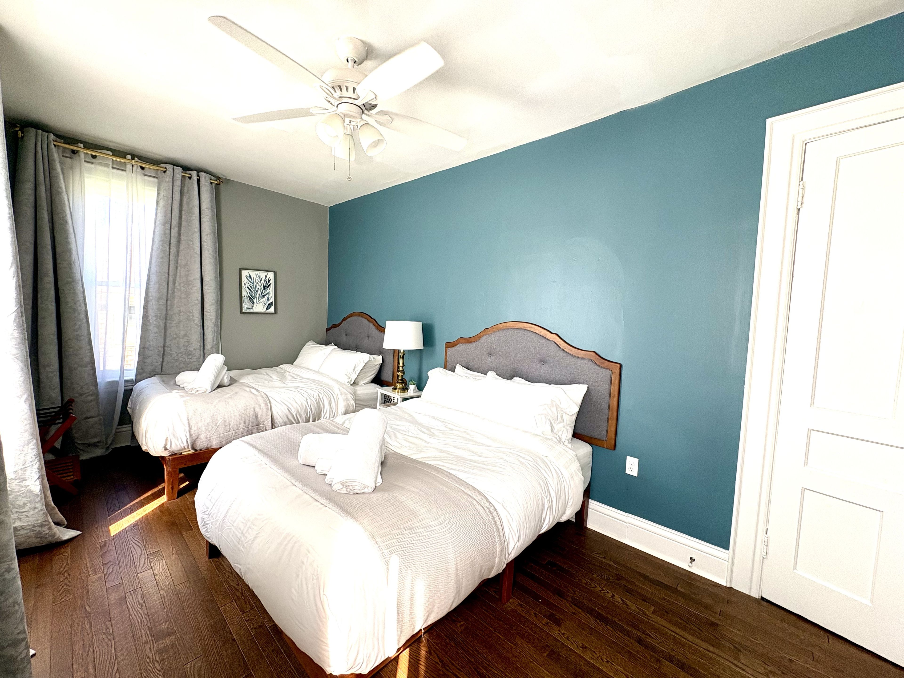 A hotel room with two beds, a ceiling fan, a nightstand with a lamp, and a door.