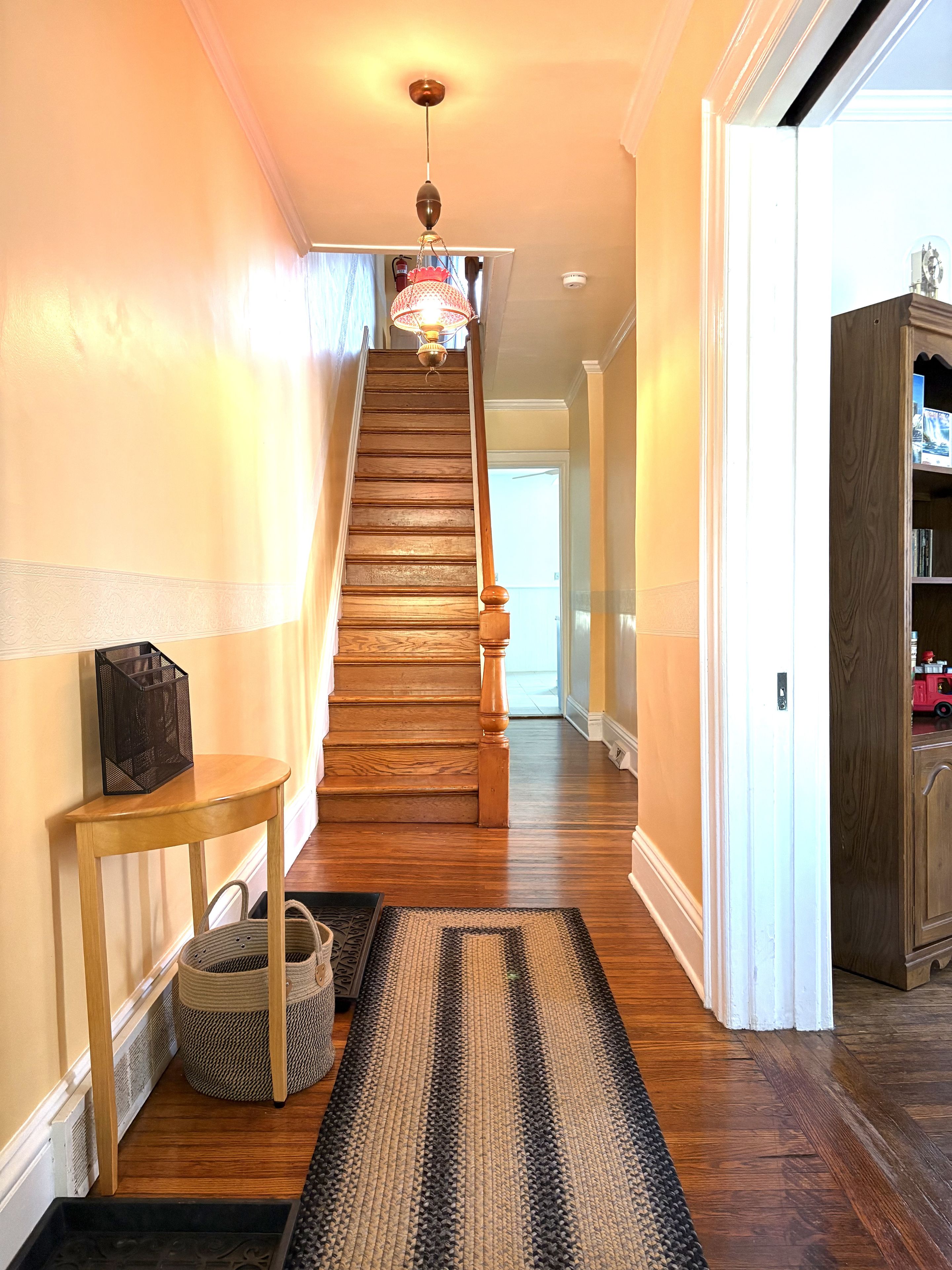 Un escalier en bois avec une rampe, un luminaire suspendu et un tapis menant à une porte.