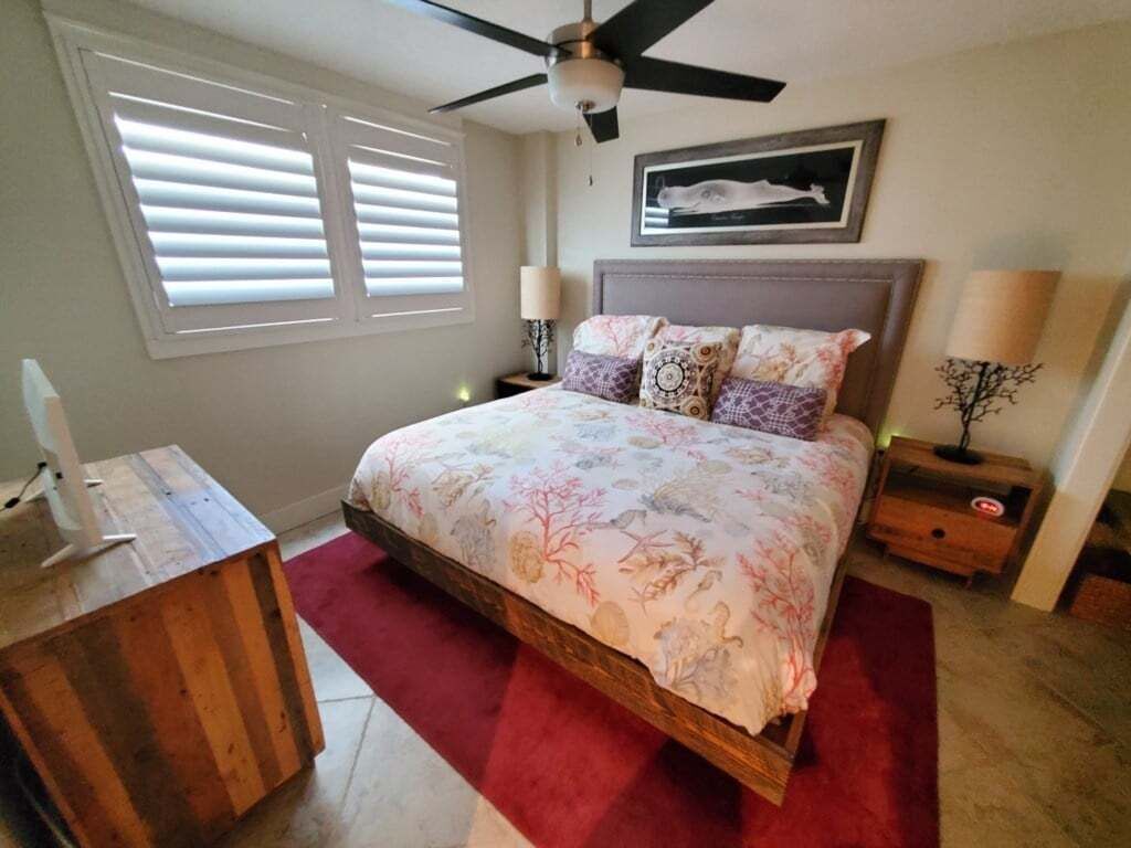 A bedroom with a bed, a ceiling fan, a nightstand, a lamp, a wooden dresser, and a window with blinds.