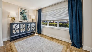 A room with a dark console table, two lamps, a painting, and a window with blinds and curtains. - 733a And B Tarpon Avenue 6 Bedroom Home (Fernandina Beach)