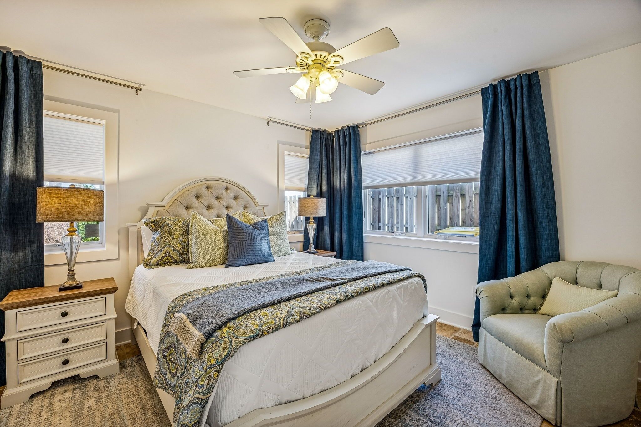 Ein Schlafzimmer mit einem großen Bett, einem Nachttisch, einem Sessel, einem Deckenventilator und einem Fenster mit Vorhängen.