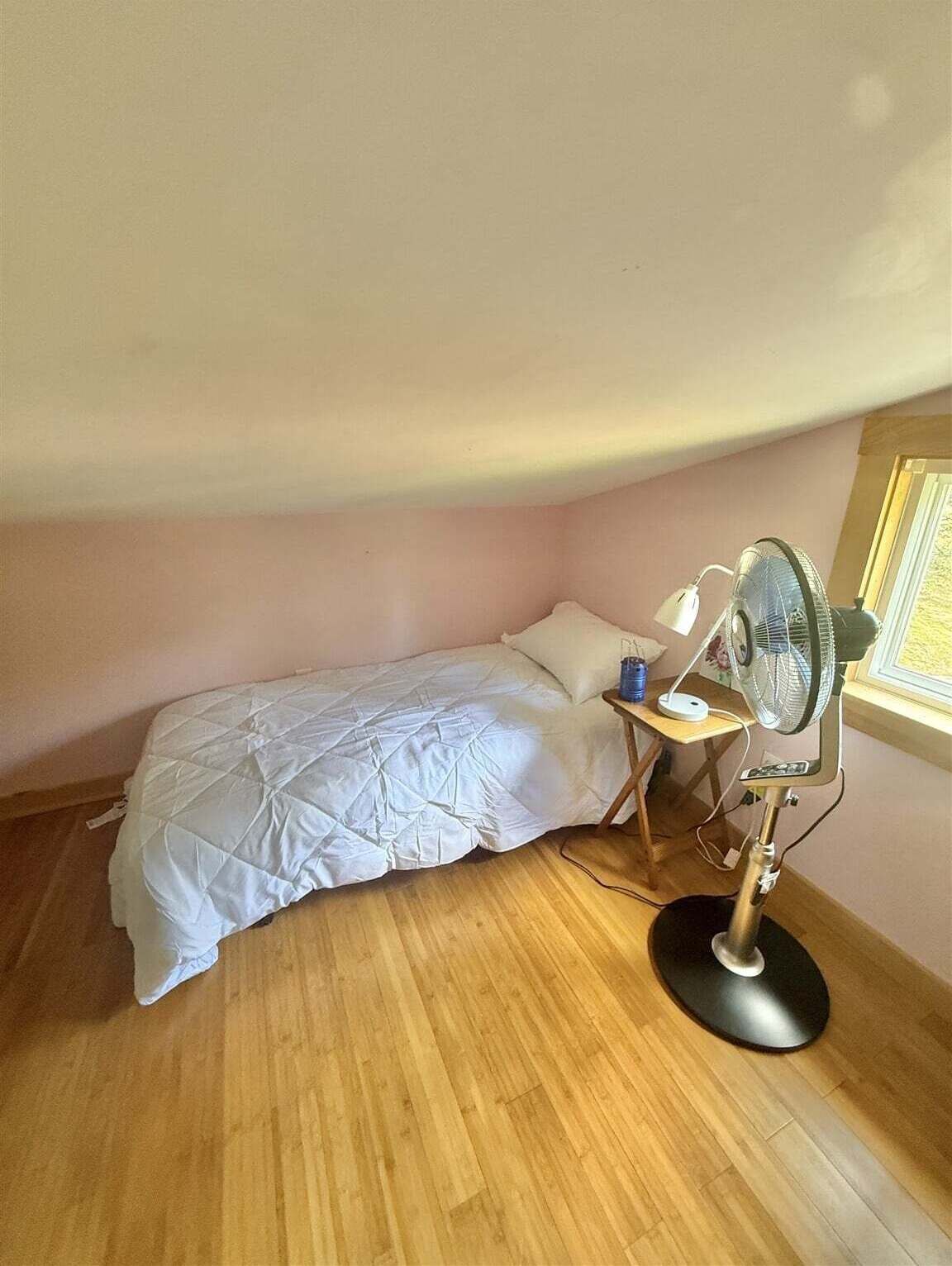 A small, single-bed room with a wooden floor, a bedside table with a lamp, and a standing fan.