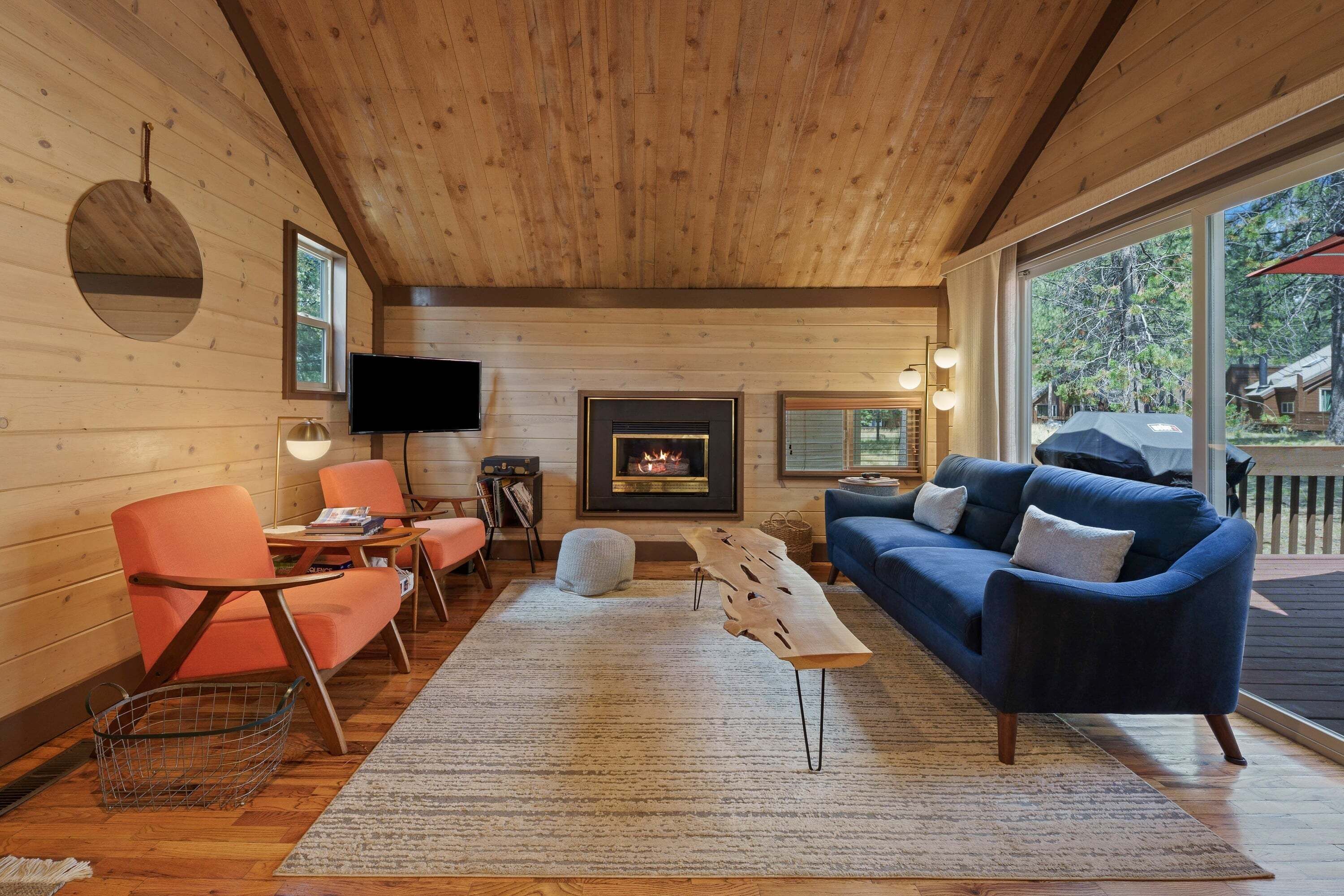 A cozy cabin interior with a fireplace, a television, and a large window offering a view of the outdoors.