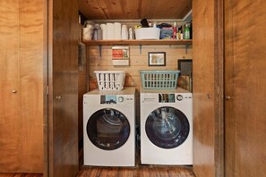 A laundry room with two front-loading washing machines, a drying rack, and shelves with various items. - 16 Pole House 2 Bedroom Home (Bend)