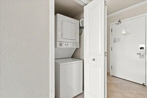 A laundry room with a washer and dryer, a door with a lock, and a wall-mounted shelf with a white object. - Maui Banyan H502 1 Bedroom Condo (Kihei)