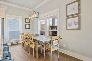 A dining area with a wooden table and chairs, framed artwork on the wall, and a view of the outdoors through the windows. - Easy Breezy 4 Bedroom Home (Port Bolivar)