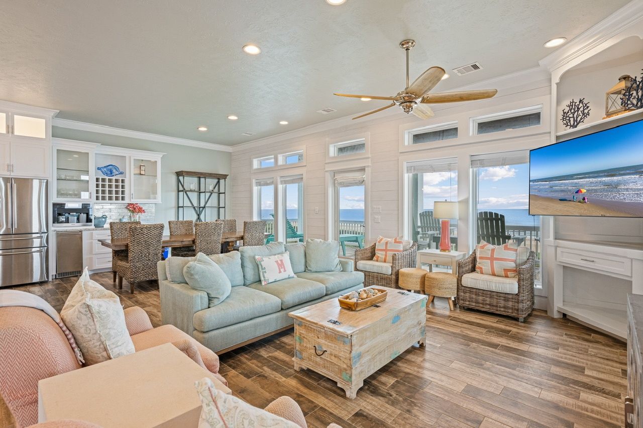 A spacious living area with a large sofa, a coffee table, and a dining table. There is a kitchen area with white cabinets and a refrigerator. The room has large windows with a view of the ocean.